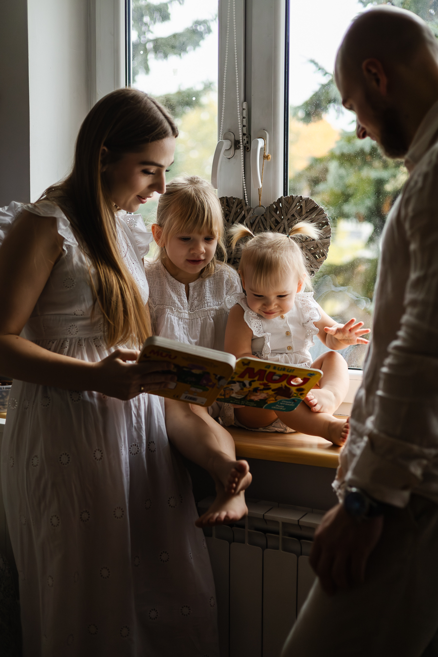 Rodzina przy oknie, mama i tata stoją, mama trzyma książkę. Na parapecie, tyłem do okna siedzą dwie małe dziewczynki i patrzą na książkę.