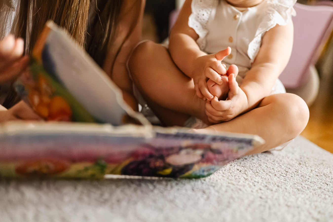 Zbliżenie na stópki i rączki małej dziewczynki, której mama czyta książeczkę.