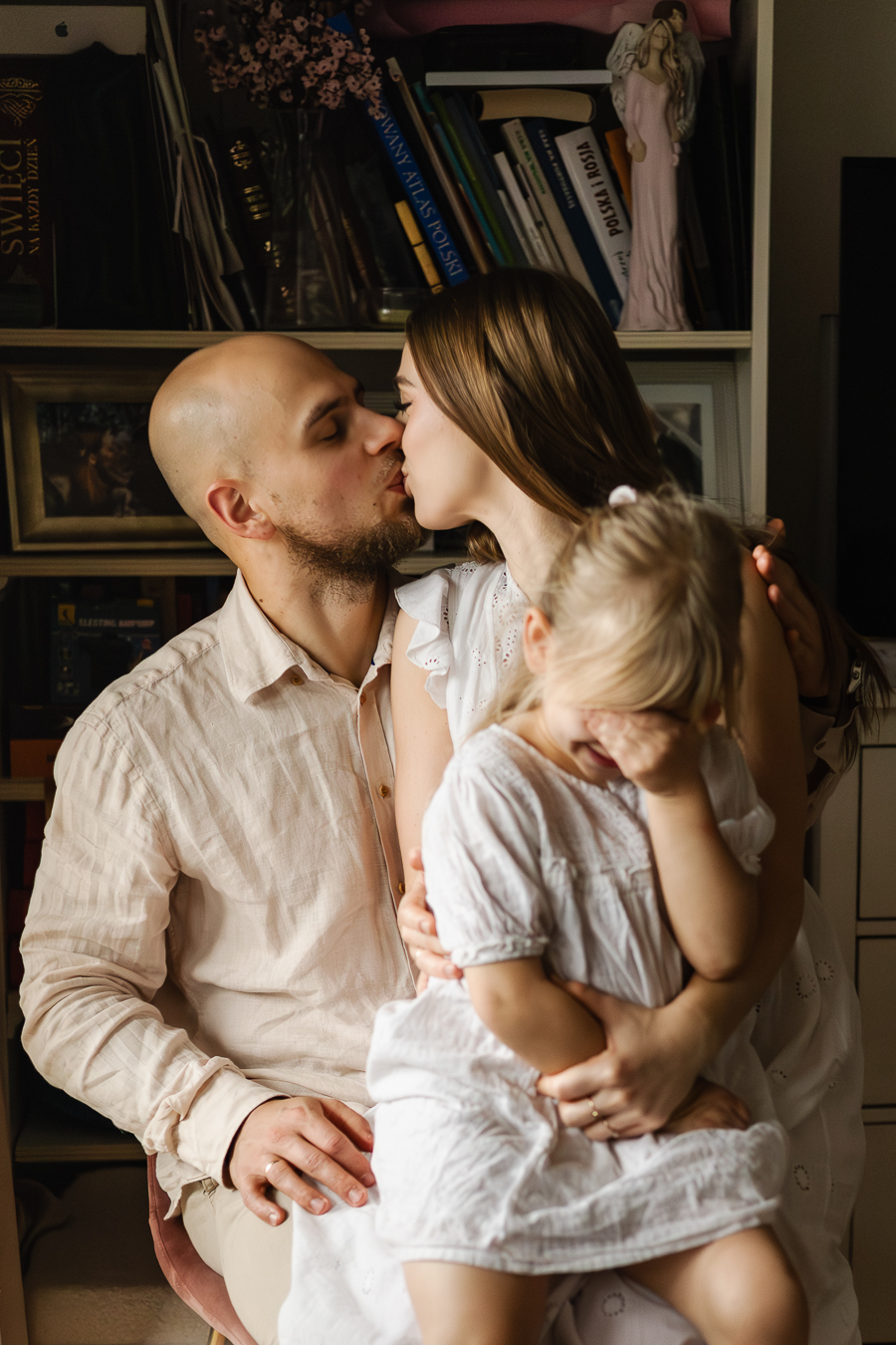 Rodzice i córka siedzą na krześle w salonie. Rodzice dają sobie buziaka a córeczka zakrywa oczy i śmieje się.