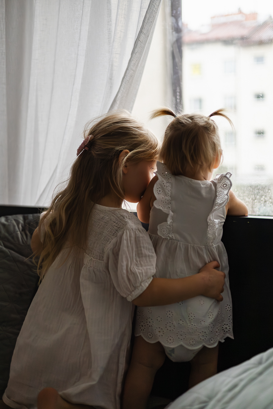 Małe dziewczynki ubrane na biało, przytulają się przy oknie, na którym są krople deszczu.