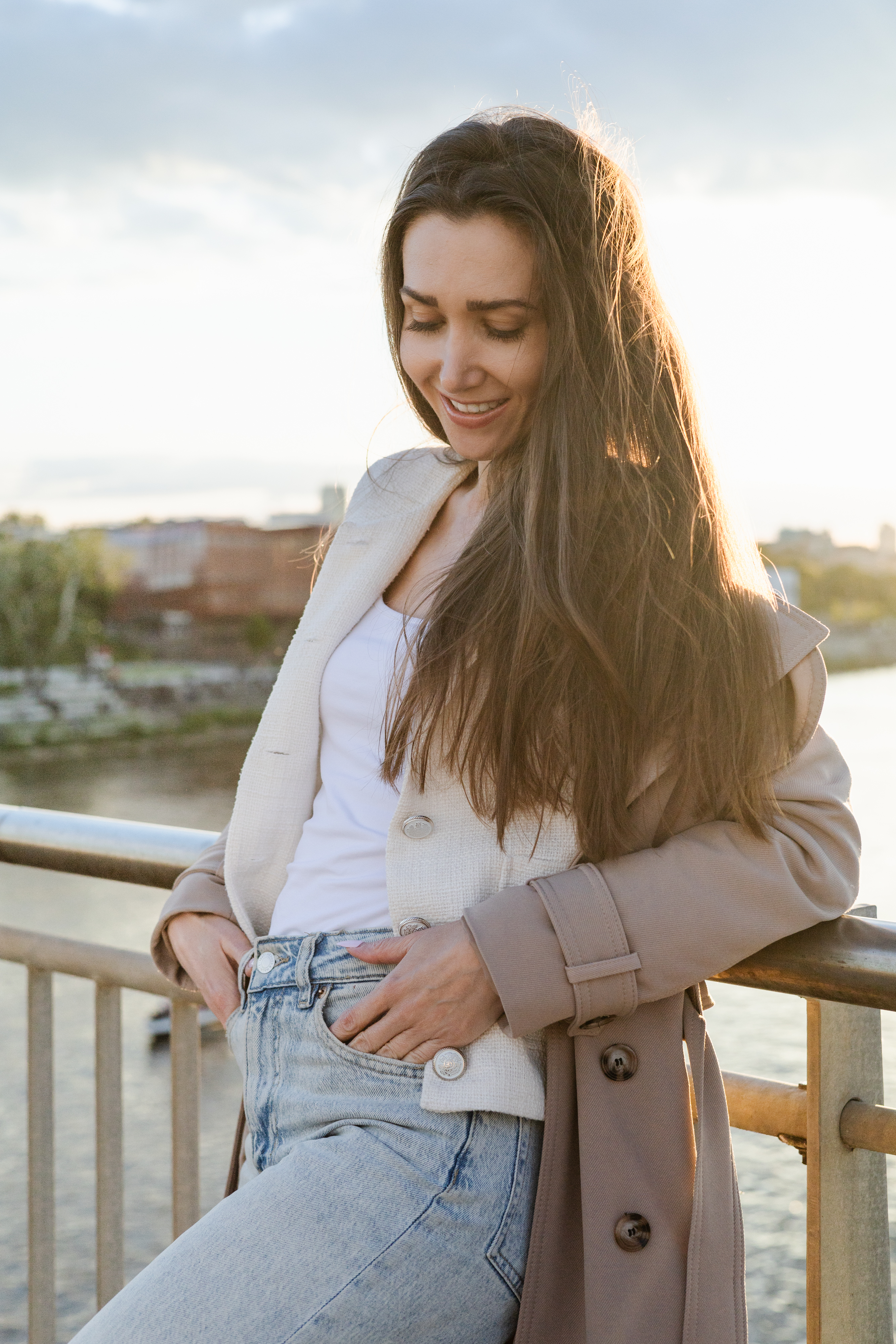 Kobieta opiera się o barierkę mostu przy rzece. Patrzy w dół i delikatnie się uśmiecha. Jej włosy oświetla zachodzące słońce.