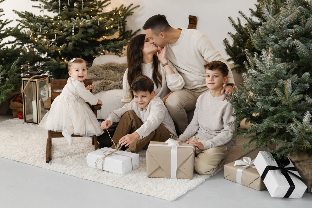 Świąteczna sesja rodzinna. Tata całuje mamę, chłopcy siedzą przy prezentach a mała dziewczynka patrzy w stronę obiektywu.