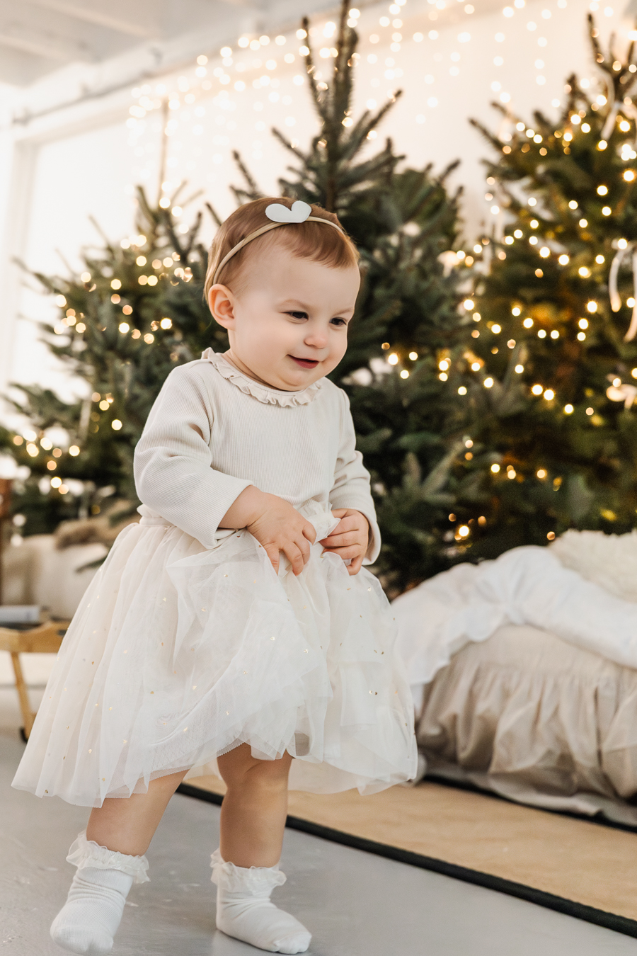Sesja świąteczna - - mała dziewczynka w białej sukience i opasce biegnie przed siebie i uśmiecha się. Za nią choinki i światełka.