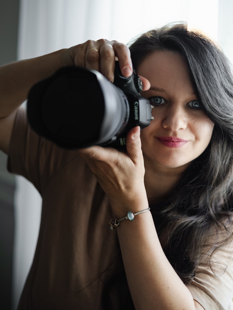 Prezentacja osoby fotografa. Fotografka z aparatem marki Canon patrzy na wprost i delikatnie się uśmiecha.