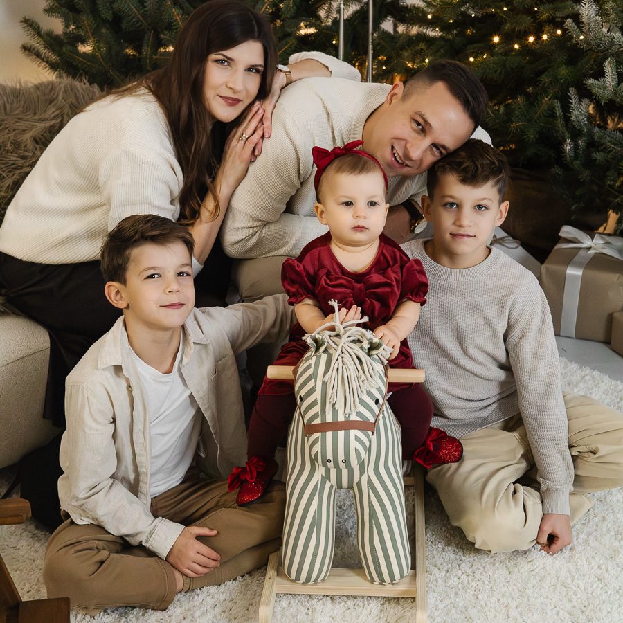 Rodzinna sesja świąteczna. Rodzina pięcioosobowa przytula się do siebie, patrzą w obiektyw. Za nimi choinki i światełka.