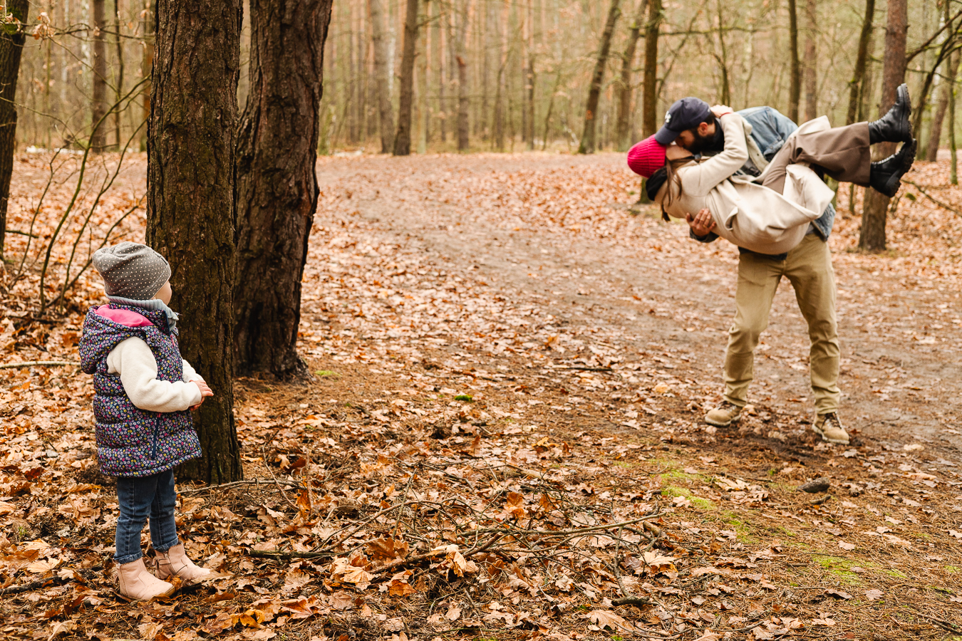 Jesienny las. Dziewczynka z za drzewa patrzy na całujących się rodziców. Tata trzyma mamę na rękach i całuje.