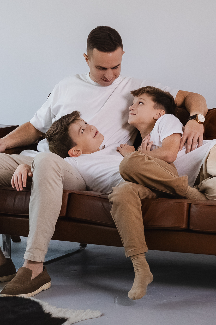 Tata i dwóch synów na kanapie - on patrzy na chłopców i uśmiecha się do nich. Chłopcy leżą i opierają głowy o tatę.